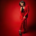 Fashion Woman in Red Pantsuit. Stylish Model with Black Bob Haircut in Eyeglasses in Trouser Suit sitting over red studio Royalty Free Stock Photo