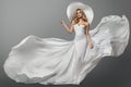 Fashion Woman in White Flying Dress over Gray Background. Elegant Bride in Wedding Gown and White Summer Hat. Beautiful Girl in Royalty Free Stock Photo