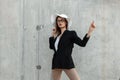 Fashion young girl in glasses in stylish business black-white clothes and beautiful straw hat stand and dancing near gray wall Royalty Free Stock Photo