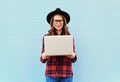 Fashion young smiling woman holding laptop computer in city, wearing a black hat, red checkered shirt over blue background Royalty Free Stock Photo