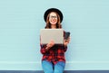 Fashion young smiling woman holding laptop computer with coffee cup resting outdoors in city, wearing black hat red checkered Royalty Free Stock Photo