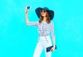 Fashion young smiling woman is taking a picture on a smartphone wearing a straw hat, white pants with a handbag clutch Royalty Free Stock Photo