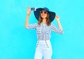Fashion young smiling woman is taking a picture on a smartphone wearing a straw summer hat, white pants over colorful blue Royalty Free Stock Photo