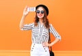 Fashion young woman model taking picture self portrait on smartphone wearing black hat white pants over colorful orange Royalty Free Stock Photo