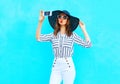 Fashion young woman is taking a picture on a smartphone wearing a straw summer hat, white pants over colorful blue Royalty Free Stock Photo