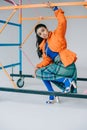 fashionable african american female model in orange winter jacket, checkered skirt and blue tights posing near colorful scaffold