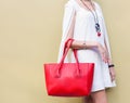 Fashionable beautiful big red handbag on the arm of the girl in a fashionable white dress, posing near the wall on a