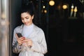 Fashionable beautiful woman with dark hair tied in pony tail dressed in white elegant coat holding cell phone looking into screen Royalty Free Stock Photo