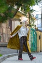 Fashionable beautiful young woman with blond hair in a stylish long coat, checkered pants, red shoes and glasses poses Royalty Free Stock Photo