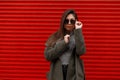 Fashionable beautiful young woman in a green fashion coat and gray sweater straightens stylish sunglasses near the red metal wall. Royalty Free Stock Photo