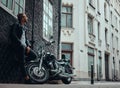 Fashionable biker dressed in a black leather jacket and jeans leaning on a wall near his retro motorcycle on old Europe