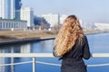 Fashionable blonde woman with long curly hair standing on the bridge and looking at the river and city Royalty Free Stock Photo