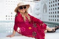 Fashionable blonde woman wearing a colorful swim coverup, sunglasses and beach hat relaxes by a resort swimming pool