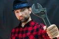 A fashionable car service employee in a red shirt and a black hat holds a large adjustable wrench in his hand on a black