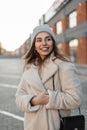 Fashionable cheerful young woman in a vintage hat in a trendy coat with a fashion black leather handbag posing and smiling in a Royalty Free Stock Photo