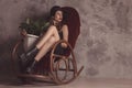 Fashionable confident woman with long legs in black dress and hat, sitting on rocking chair posing in studio with beton background Royalty Free Stock Photo