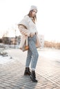 Fashionable cute young woman in a stylish eco coat in a knitted hat in blue jeans in trendy leather black boots posing outdoors on