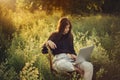 Fashionable elegant girl working on laptop and sitting on rustic chair in warm sunshine in summer meadow at sunset. Young business Royalty Free Stock Photo