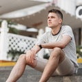 Fashionable European young man in a stylish striped t-shirt in summer beige shorts sits on the stairs on the street Royalty Free Stock Photo