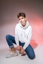 Fashionable handsome young European male model dressed in white sweatshirt, blue jeans and white sneakers posing in studio on a Royalty Free Stock Photo