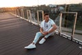 Fashionable handsome young man in stylish clothes in sneakers resting on the floor near a glass wall at sunset in the city. Royalty Free Stock Photo