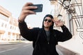 Fashionable happy hipster man in dark sunglasses in stylish hooded sweatshirt makes selfie on phone while standing in the city Royalty Free Stock Photo