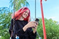 Fashionable happy teenage girl with dyed hair with smartphone riding swing