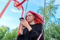 Fashionable happy teenage girl with dyed hair with smartphone riding swing