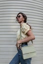Fashionable hipster curly woman in vintage round sunglasses in a yellow blouse top in stylish blue jeans with a handbag walks near Royalty Free Stock Photo