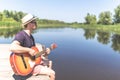 Fashionable hipster guy sitting on wooden dock and holding acoustic guitar against beautiful lake Royalty Free Stock Photo