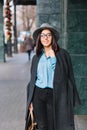 Fashionable joyful young business woman in black glasses walking on street in big city. Blue shirt, grey coat, hat Royalty Free Stock Photo