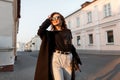 Fashionable joyful young hipster woman in a long coat in dark sunglasses in a t-shirt in white jeans with a leather bag