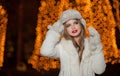 Fashionable lady wearing white fur accessories outdoor with bright Xmas lights in background. Portrait of young beautiful woman Royalty Free Stock Photo