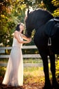 Fashionable lady with white bridal dress near brown horse in nature. Beautiful young woman in a long dress posing with a horse