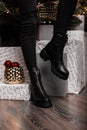 Fashionable leather black women`s winter boots. Young woman in jeans in stylish shoes stands in a room near the holiday boxes and