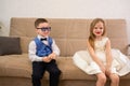 A fashionable little boy in a business suit and glasses and a girl in a white dress and beads are sitting on the sofa Royalty Free Stock Photo