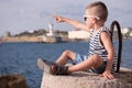 Fashionable little boy in sunglasses and vest points finger into distance sitting against sea