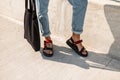 Fashionable men`s legs in vintage jeans in trendy red leather sandals with a black fabric bag stand on the pavement.