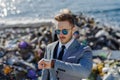 Businessman looking at watch, checking time standing on pile of waste on beach. Consumerism versus pollution concept Royalty Free Stock Photo