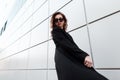 Fashionable modern hipster young woman in a black stylish coat in vintage dark sunglasses in posing near a modern building Royalty Free Stock Photo