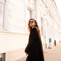 Fashionable modern young brunette hipster woman in black elegant long coat in stylish sunglasses posing near a white building Royalty Free Stock Photo