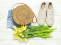 Fashionable natural organic round rattan bag, denim shorts, beige women`s espadrilles, yellow narcissus daffodil flowers on light