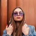 Fashionable portrait American young woman in a stylish blue denim jacket in trendy glamorous purple glasses near a metallic modern Royalty Free Stock Photo