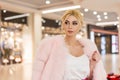 Fashionable portrait pretty luxurious young woman with blond hair in chic pink expensive fur coat in elegant white top in shopping