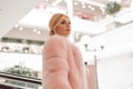 Fashionable portrait pretty luxurious young woman with blond hair in chic pink expensive fur coat in shopping center. Beautiful