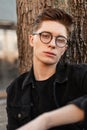 Fashionable portrait serious sexy young man hipster with trendy hairstyle in stylish glasses in vintage black denim jacket near Royalty Free Stock Photo