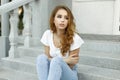 Fashionable pretty european young woman in a white stylish t-shirt in blue fashionable jeans is resting on the vintage stair