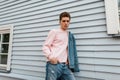 Fashionable pretty young man in pink stylish sweatshirt in denim blue jacket in jeans stands near white vintage building on street Royalty Free Stock Photo