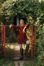 A fashionable rural woman in a red dress and high boots stands leaning on a red fence.