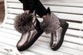 Fashionable silver shoes with gray fur on female legs. Stylish young woman in a glamorous shiny winter shoes sits on a wooden